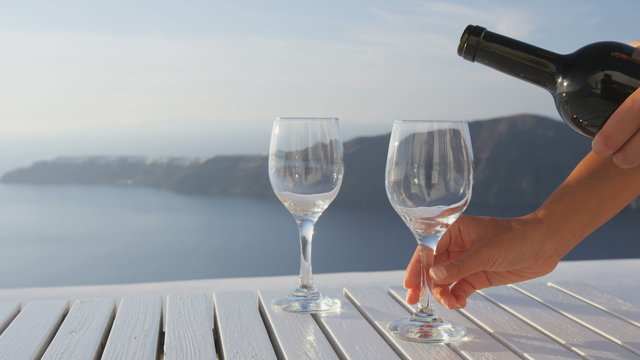 Woman serving red wine from bottle outdoors in beautiful nature. Hands of woman pouring red wine in glass on table. Female is serving alcohol on luxury vacation travel on Santorini, Greece. RED EPIC.
