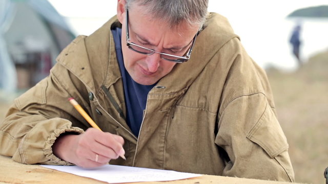 Man poet or a writer writes a new work, verse or poem. Maybe wrote a letter to his wife or family. It looks like a fisherman or a hunter. The glasses and a wooden table