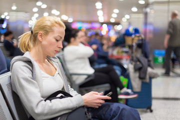 Casual blond young woman using her cell phone while waiting to board a plane at the departure gates. Wireless network hotspot enabling people to access internet conection. Public transport.