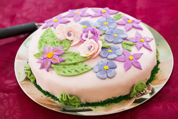 Праздничный торт в пастельный тонах, украшенный нежно-розовой кондитерской мастикой и кремовыми цветами  фиолетового, голубого, розового и зеленого цветов 
