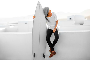 Young smiling woman in white t-shirt and hat sitting with surfboard on the roof top on the white city background in the morning