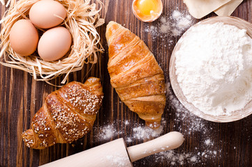 Ingredients for baking croissants - paper, flour, wooden spoon, rolling pin, eggs, egg yolks, butter served on a rustic wooden tray table.