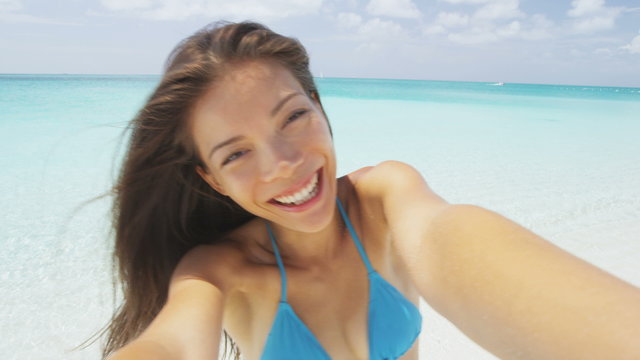 Selfie on beach travel. Excited young woman taking self-portrait on having fun. Carefree girl in 20s is enjoying her summer vacation. Portrait of happy female in blue bikini top smiling joyful.