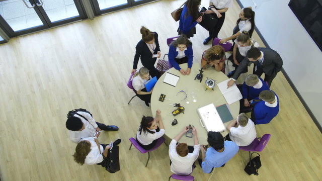 View from above of students and teachers in a technology lesson. Some are sitting at the table whilst others are standing and talking amongst themselves.