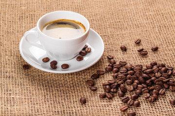 Beautiful white coffee cup and saucer and coffee beans on the background of burlap. Dark background.