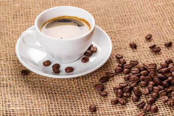 Beautiful white coffee cup and saucer and coffee beans on the background of burlap. Dark background.