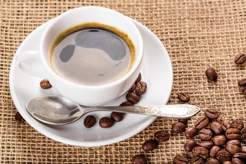 Beautiful white coffee cup and saucer and coffee beans on the background of burlap. Dark background.