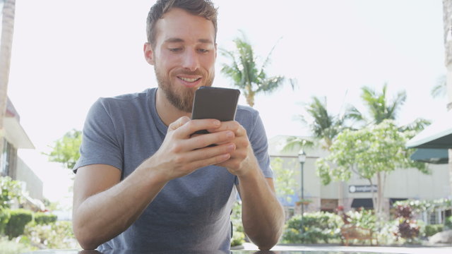 Smiling young man using mobile smart phone at sidewalk cafe. Bearded male is text messaging on smartphone. Man in casuals is sitting at outdoor restaurant table. RED EPIC.