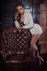 beautiful slim girl in stylish fashion short white shorts and a jacket is leaning on a big brutal leather brown armchair