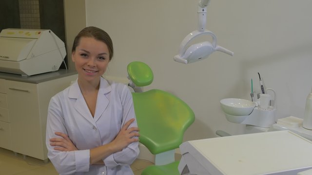 Dentist Young Female Doctor in Lab Coat Smiling Sitting on a Chair in a Ward Looking at Camera Dentist's Machine Dental Lamp Green Chair Dental Clinic