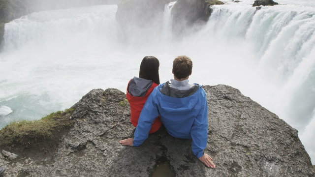 Young couple watching Godafoss waterfall from cliff. Man and woman are relaxing on rock during vacation. Male and female are enjoying majestic view of famous attraction in Iceland.