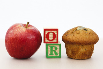 Rosy red apple and a muffin cake with the word OR on a tabletop to illustrate choice.