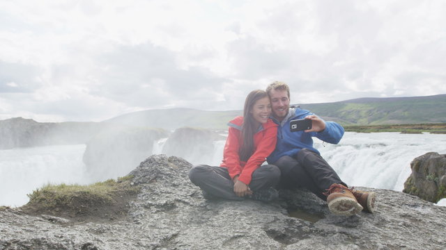 Cheerful couple taking selfie against majestic Godafoss waterfall. Happy male and female tourists are sitting on cliff. Man and woman are visiting famous tourist attraction of Iceland.