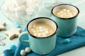 Two mugs of hot cacao with marshmallow, cookies and napkin on blue table