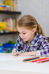 Cute smiling little girl with blond hair sitting at white table and drawing with multicolored pencils