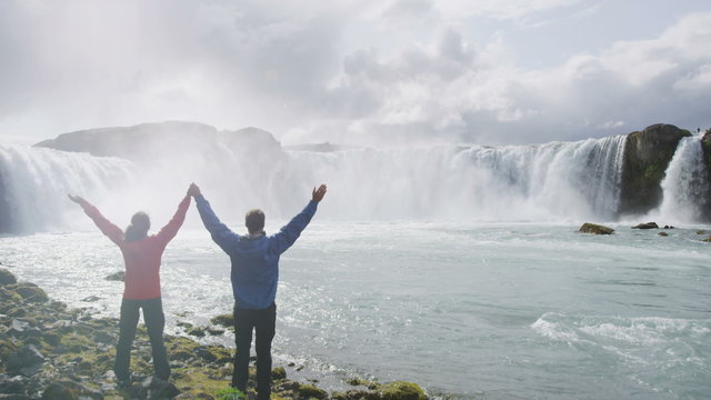 Carefree multiethnic couple raising hands at spectacular Godafoss waterfall. Happy tourists are visiting famous attractions. Man and woman are enjoying vacation at beautiful waterfall in Iceland.