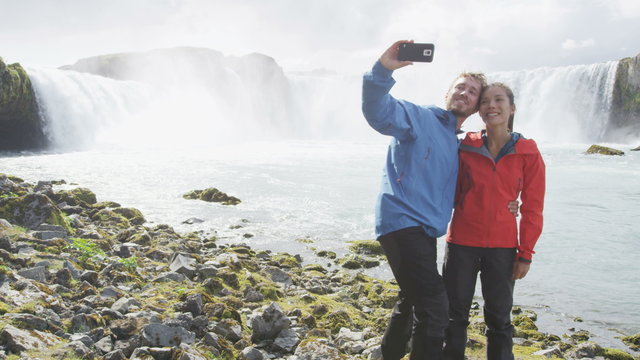 Smiling young couple photographing against spectacular Godafoss waterfall. Male and female tourists are taking selfie during vacation at Iceland. Man and woman are visiting famous tourist attractions.