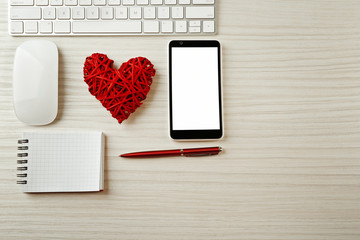 Computer peripherals with wicker red heart, notebook and mobile phone on light wooden table