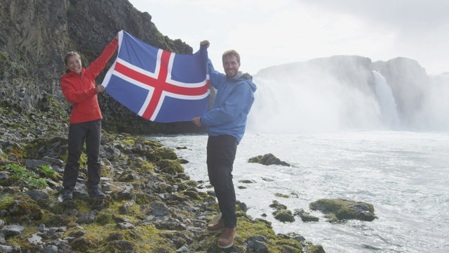 Excited young couple holding Iceland flag against Godafoss waterfall. Portrait of happy tourist couple enjoying vacation at beautiful waterfall. Male and female are visiting famous attractions.