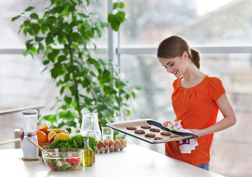 Young woman with pan of chocolate cookies near the table in kitchen