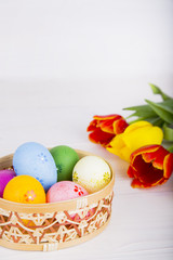 Colorful Easter eggs in wicker basket with tulips on white woode