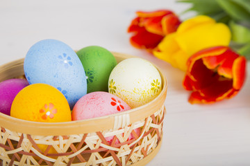 Colorful Easter eggs in wicker basket with tulips on white woode
