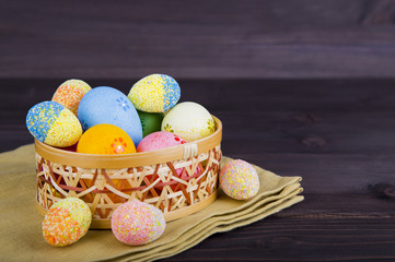 Colorful Easter eggs in wicker basket on textile on dark wooden