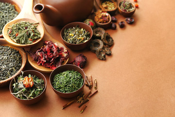 Variety of dry tea in bowls with teapot on light brown background, closeup