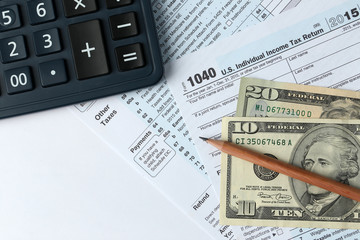 1040 Individual Income Tax Return Form for 2015 year with a pencil to fill in, calculator and dollar bills on the white desk, top view