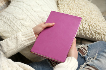 Young woman sitting on the sofa with white cushions and holding a purple book cover, close up