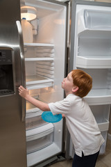 Hungry little boy looks sadly into an empty refrigerator in a poor middle-class home.