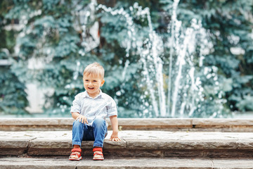 Portrait of cute adorable funny Caucasian  little boy toddler in white shirt and blue jeans playing laughing smiling having fun by fountain in summer outside