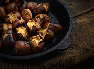 Heap of grilled edible chestnuts in cast iron skillet over dark wooden surface with textile napkin
