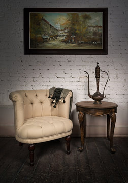 Vintage still life of old beige armchair, brass teapot, hanged framed painting and two small antique nested tables on dark brown wooden floor and white bricks wall in Studio