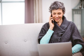 Mature woman with laptop talking on mobile phone at home