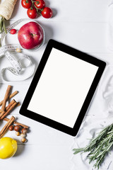 Tablet with a measuring tape, rosemary, nuts, parsley root, tomatoes, cinnamon and red apples on a white wooden background top view vertical, space for publicity information or advertising text