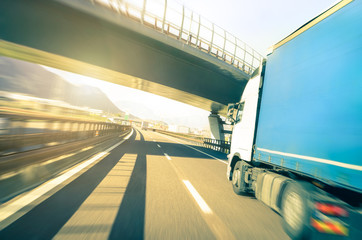 Generic semi truck speeding on highway under overpass - Transport industry logistic concept with semitruck container driving fast on speedway - Soft vintage filter with sunshine halo and blurred edges - obrazy, fototapety, plakaty