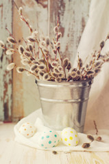 Easter eggs with a bouquet of spring branches of a willow in a bucket on a wooden background.