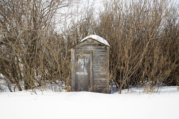 A small gray wooden weathered outhouse surrounded by bare trees in winter
