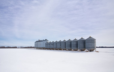 A row of steel industrial grain storage bins in a winter countryside landscape