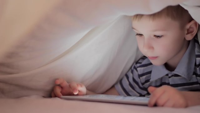Boy lying under blanket and watching cartoon on touch pad