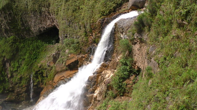 The powerful Agoyan waterfall plunges down a rugged moss-covered cliffside surrounded by dense tropical vegetation near Banos, Ecuador, the camera tilting down to reveal the turbulent river below.