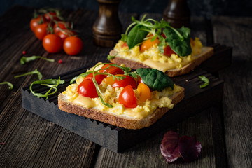 Breakfast toast with scrambled eggs, cheese, cherry tomatoes, arugula and corn salad on rustic wooden background