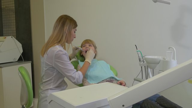 Woman Dentist is Examining a Teeth With Mirror Patient Teenage Girl is Sitting in a Green Chair Woman Doctor Puts an Instruments Shifts the Table Clinic