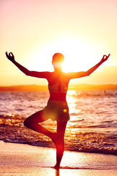 Wellness of mind - Yoga woman standing on one leg doing tree pose with open raised arms in sunset flare in front of the ocean on beach. Mindfulness and meditation concept.
