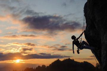 Silhouette of beautiful athletic woman climbing steep rock wall against amazing sunset scene in the mountains. Girl is hanging on one hand and looking up. - obrazy, fototapety, plakaty