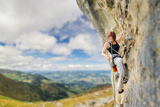 Portrait of young female rock climber on steep overhanging rock cliff. Holding the rope and looking down. Beautiful blue sky and mountains on the background.
