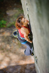 Young beautiful girl climber climbing steep boulder, searching for next grip. Top view on focused muscular female athlete. Climbing equipment.