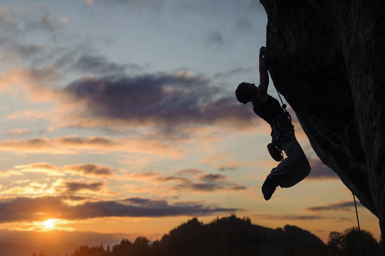 Silhouette of athletic woman rock climber climbing steep rock wall against amazing sunset sky in the mountains. Girl is hanging on two hands.