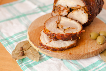 Close up shot of sliced pork meatloaf on wooden cutting board on white and green towel on light wooden background with bay leaves, black pepper and olives